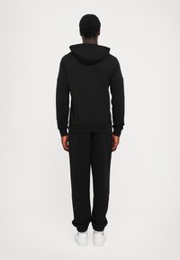Personne portant un sweat à capuche noir et un pantalon de survêtement noir, de dos face au spectateur, baskets blanches visibles.