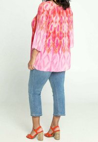 Donna che indossa una blusa a fantasia rosa e rossa, jeans a vita alta blu e sandali con tacco arancioni e zeppa in sughero, in piedi di lato.