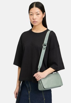 Frau trägt ein lockeres schwarzes Oberteil und einen dunklen Jeansrock, mit einer hellgrünen Umhängetasche mit breitem verstellbarem Schulterriemen über der Schulter.