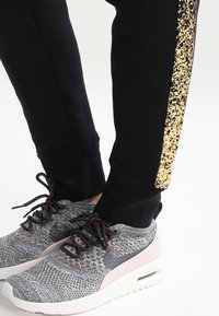 Svarta träningsbyxor med en guldprickig sidremsa, kombinerade med grå stickade sneakers som har en rosa detalj och vit sula.