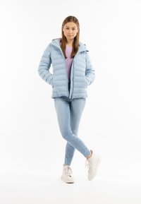 Giacca a vento azzurra chiara con cappuccio, design trapuntato, zip frontale, indossata sopra un maglione lilla e jeans azzurri chiari, abbinata a sneakers bianche.