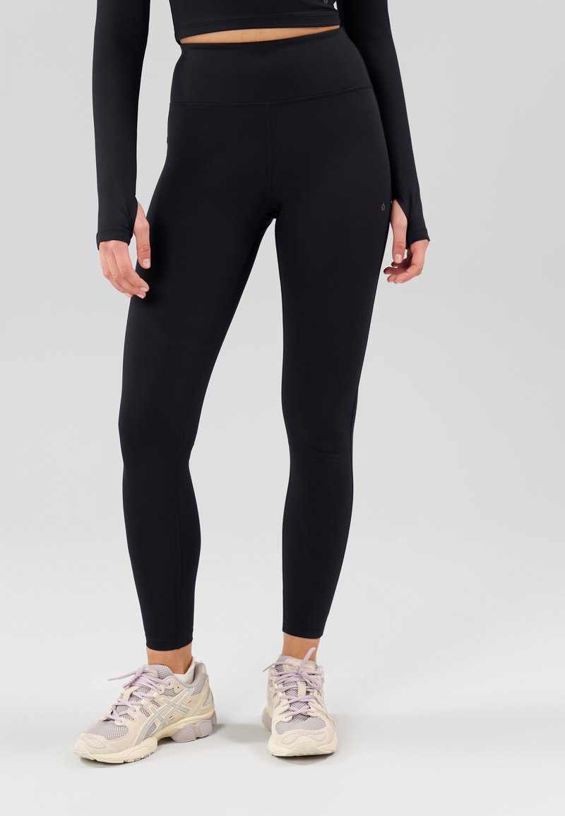 Schwarze Leggings mit hoher Taille und glatter Textur, nahtlos gestaltet. Kombiniert mit hellfarbigen Sportschuhen.