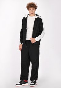 Sort zip-up jakke med hvide accenter, båret over en hvid t-shirt. Sorte løse bukser og sort-røde sneakers fuldender outfittet.