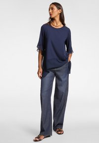 Top blu navy con maniche trasparenti e svasate, abbinato a pantaloni in denim chiaro e comodi. Il modello è in una posa rilassata.