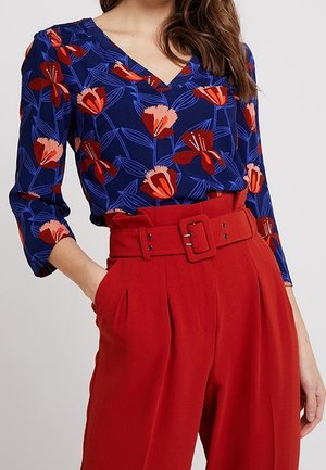 Kobieta ubrana w niebieską bluzkę w kwiaty z dekoltem w serek i rękawami do łokcia, włożoną do wysokich czerwonych spodni z szerokim paskiem i klamrą.