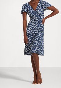 Blå blommig omlottklänning med korta volangärmar, V-ringning och en utsvängd kjol. Har vita bladmönster och en mjuk tygtextur. Bruna klacksandaler.