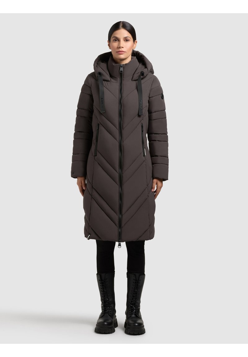 Langer, dunkelgrauer Puffercoat mit Chevron-Muster, Kapuze, zwei seitlichen Reißverschlusstaschen und schwarzem Zubehör. Getragen mit schwarzen Hosen und Stiefeln.