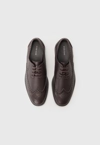 Zapatos de vestir de cuero marrón con diseño tipo wingtip, detalles perforados y cordones redondos. La suela es plana con un ligero aumento de altura.