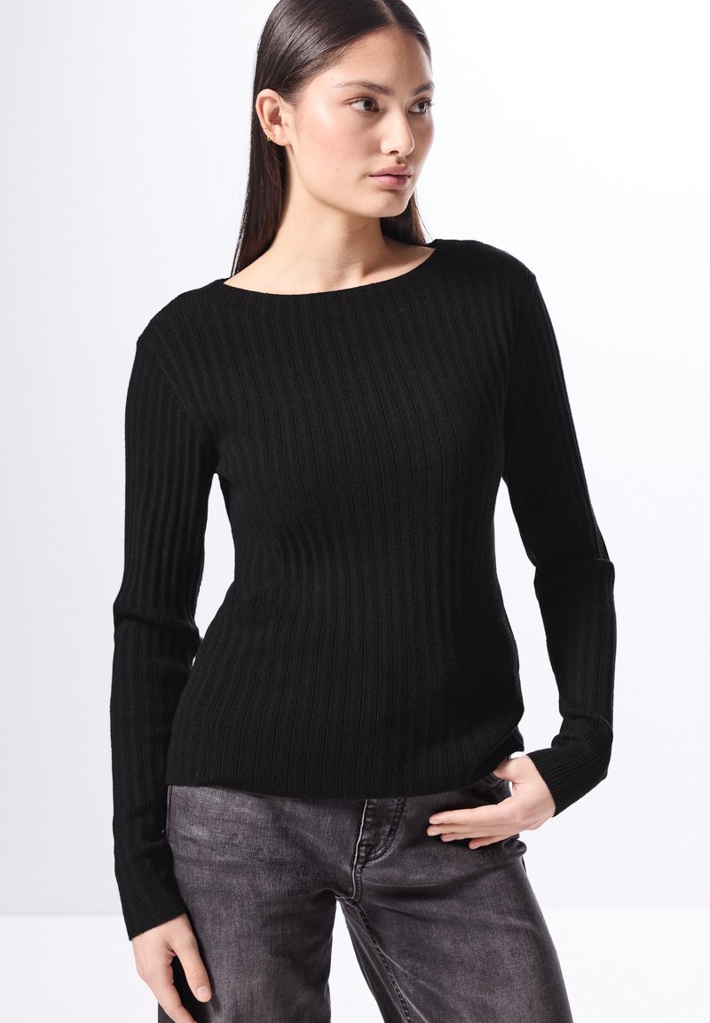 Maglione nero a coste con scollatura a barca e maniche lunghe, abbinato a jeans grigio scuro, situato su uno sfondo luminoso.