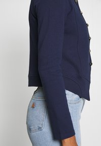 Personne portant un cardigan bleu marine à manches longues boutonné et un jean en denim bleu clair, vue de profil sur un fond uni.