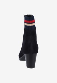 Botín de ante azul marino con un tacón ancho y un puño de punto con rayas rojas, blancas y azules. Textura suave con un diseño ajustado.