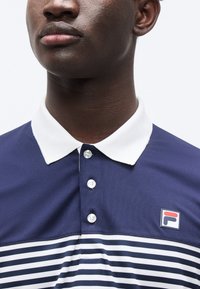 Σκούρο μπλε μπλούζα πόλο με λευκό γιακά και κουμπιά, με οριζόντιες λευκές και μπλε ρίγες στο κάτω μέρος. Λογότυπο στο στήθος.