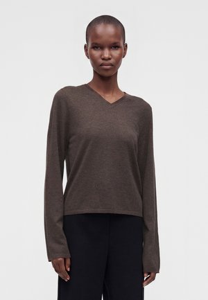 Kvinde med kort hår bærer en brun langærmet V-hals sweater og sorte bukser, stående mod en ensartet lys baggrund.