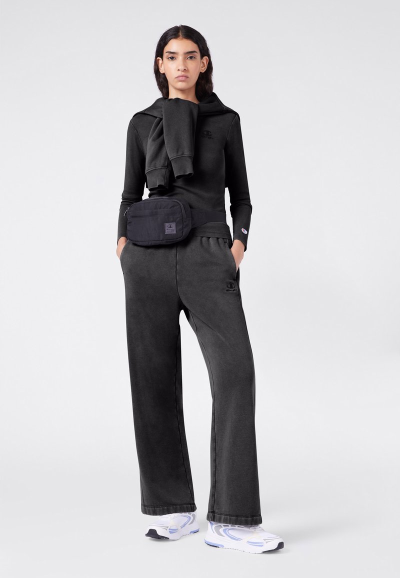 Młoda osoba nosząca czarną bluzę z kapturem, z bluzą narzuconą na ramiona, czarne szerokie spodnie, czarną saszetkę w pasie i białe trampki.