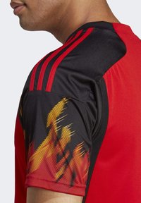 Jersey deportivo rojo y negro con tres franjas rojas en el hombro y un patrón de llamas en amarillo y negro en la manga.