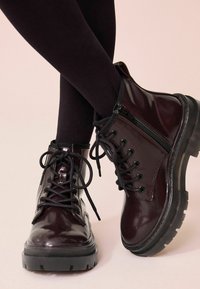 Bottes en cuir verni bordeaux avec une semelle en caoutchouc noire, dotées de fermeture à lacets et à zip sur le côté, d'accents texturés et d'un design à bout rond.
