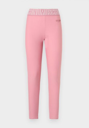 Vrouw die blootsvoets staat, draagt een roze sportbeha en leggings met "MOSCHINO" gedrukt op de tailleband tegen een witte achtergrond.