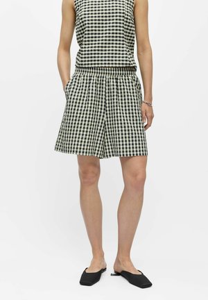 Nő fekete-fehér kockás ujjatlan felsőt és hozzáillő térdig érő rövidnadrágot visel fekete bebújós cipővel, zsebre tett kézzel áll.