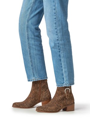 Enkelhoge laarzen met luipaardprint in bruin suède, voorzien van een gebogen hak en decoratief metalen beslag, gecombineerd met lichtblauwe spijkerbroeken.