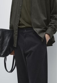 Donkerblauwe broek van een katoenmengsel, met een knoop in de tailleband en zijzakken, gecombineerd met een donkergroen shirt en een zwarte leren tas.