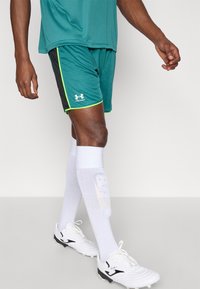 Zaļgani zilas sporta šortos ar melnām sānu panelēm un zaļu apmali, kombinēti ar baltām ceļgalu zeķēm un baltām futbola kurpēm ar sportisku dizainu.