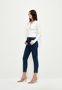 Bílý žebrovaný kardigan s zapínáním na knoflíky, kombinovaný s tmavě modrými džínami s vysokým pasem a béžovými lodičkami na podpatku, předvádějící úzký siluetu.