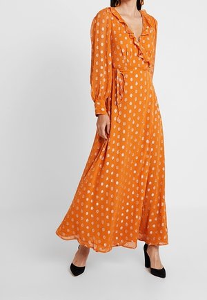 Kobieta ubrana w pomarańczową sukienkę maxi z długim rękawem, białe kropki, falbaniasty dekolt, czarne szpilki, stojąca na białym tle.