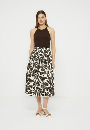 Γυναίκα όρθια, φορώντας ένα μαύρο αμάνικο τοπ και λευκή midi φούστα με ψηλή μέση και μεγάλο μαύρο λουλουδάτο σχέδιο, καθώς και λευκά σανδάλια.