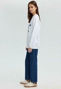 Witte sweatshirt met marineblauwe tekst, gecombineerd met blauwe rechte jeans en beige sneakers. De stof lijkt zacht en heeft een relaxte fit.