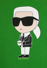 T-shirt gráfica verde com uma figura de cartoon vestindo um fato preto, camisa branca, grandes óculos de sol pretos e segurando uma chávena branca com detalhe verde.