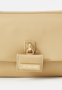Beige faux leather handbag with a gold-tone hardware accent, featuring a flap closure and a logo-embellished front pocket.