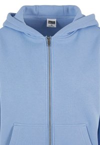 Felpa con zip di colore azzurro chiaro, dotata di cappuccio, tasche frontali e materiale morbido in misto cotone. Presenta una zip argentata e un'etichetta con il logo all'interno del colletto.