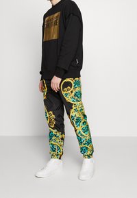 Černá mikina se zlatým logem na hrudi, kombinovaná s vícebarevnými kalhotami s potiskem leopardího vzoru a zlatými akcenty. B�ílé tenisky.