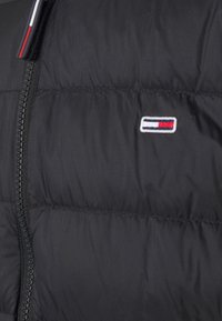 Mustat puffertiliivi, jossa on vetoketjullinen etuosa ja pieni brodattu logo punaisena, valkoisena ja sinisenä vasemmassa rintamuksessa. Sileä pinta.