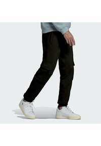 Pantalones deportivos negros con bolsillos tipo cargo, puños acanalados y cinturilla ajustable, combinados con zapatillas deportivas blancas. La tela parece suave y texturizada.