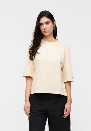 Frau mit langen dunklen Haaren, die ein beiges oversized Tommy Hilfiger T-Shirt und schwarze Hose trägt, steht vor einem schlichten hellen Hintergrund.