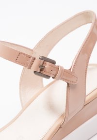 Sandale en cuir verni beige avec boucle métallique réglable et bride sur une semelle intérieure blanche rembourrée.