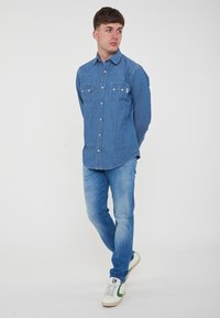 Camicia in denim blu con due tasche sul petto, collare a punta e chiusura con bottoni. Jeans azzurri e sneakers bianche con dettagli verdi.