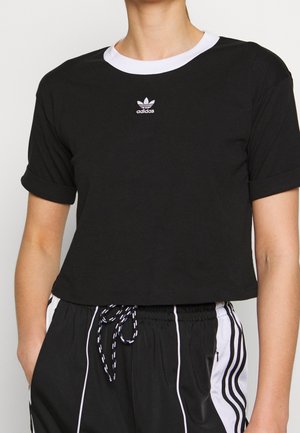 Osoba nosząca czarną krótką koszulkę Adidas z białym kołnierzykiem oraz czarne spodnie dresowe Adidas z białymi paskami i ściągaczem w talii.
