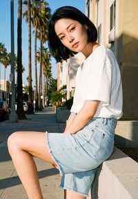 Jeune femme aux cheveux courts noirs portant un t-shirt blanc et une jupe en jean clair, assise sur un mur en béton dans une rue ensoleillée bordée de palmiers.