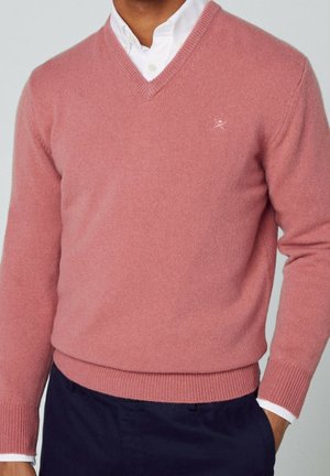 Uomo con maglione rosa a scollo a V sopra una camicia bianca abbottonata, con pantaloni scuri, in piedi con una mano in tasca.