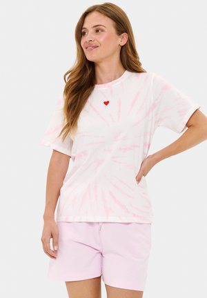 Bílý tričko s tye-dye vzorem a růžovým spirálovým motivem, s krátkými rukávy, se malým červeným srdcem na hrudi. Doplněno světle růžovými šortkami.