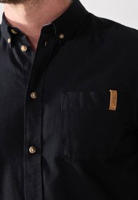 Camicia nera con bottoni, munita di taschino sul petto e etichetta in pelle beige, con bottoni color oro e una finitura in tessuto testurizzato.
