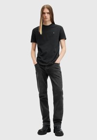 Sort bomulds T-shirt med rund hals og lille logo, parret med mørkegrå denimjeans og sorte læderstøvler. Ensfarvet baggrund.