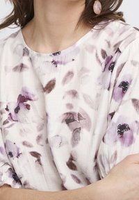 Blusa floreale in tessuto leggero con base crema e motivi di fiori viola pastello e marrone. Collo rotondo e maniche arricciate.