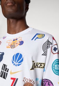 Személy fehér inget visel, amelyen színes NBA csapatlogók és kosárlabda grafikák láthatók, többek között a Lakers, Pistons és Pacers logói.