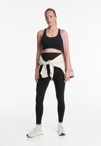 Fekete sportmelltartó és leggings szett, krémszínű pulóver a derék körül megkötve, bézs sportcipők, világos hajú modell, rózsaszín táska a vállon.