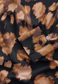 Tissu noir avec des taches abstraites marron et beige, présentant un poignet avec deux boutons noirs et un détail de manche froncée.