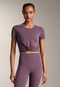 Tricou scurt de culoare mov, cu un nod în față, fabricat dintr-un material moale și elastic, purtat împreună cu leggings asortați, cu talie înaltă. Textură netedă.