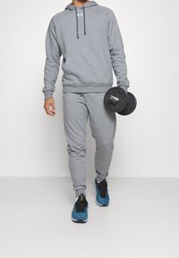 Felpa grigia con cappuccio, dotata di cordini e polsini a coste, abbinata a pantaloni della tuta coordinati. Indossata con scarpe da ginnastica blu, mentre tiene in mano un disco per il sollevamento pesi.